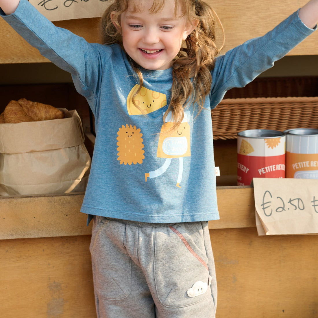 Nest Designs Munchies Long Sleeve Tee in Oceanview blue, front view on a smiling child with arms raised at a market