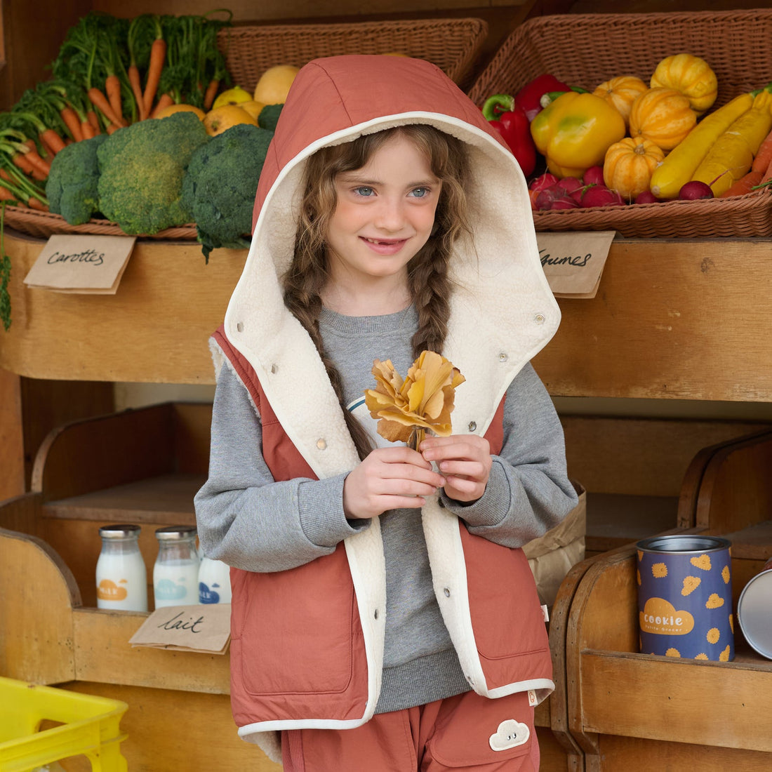 Child smiles in the Nest Designs Redwood Burl Cozy Crumbs Reversible Fleece Vest worn open with its cream-lined hood raised