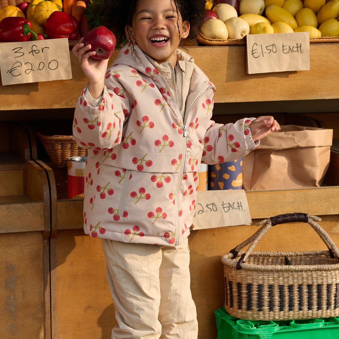 Child wears the Nest Designs You're So Vine Waterproof Down Jacket at a market while holding a red apple beside the cherry-print shell