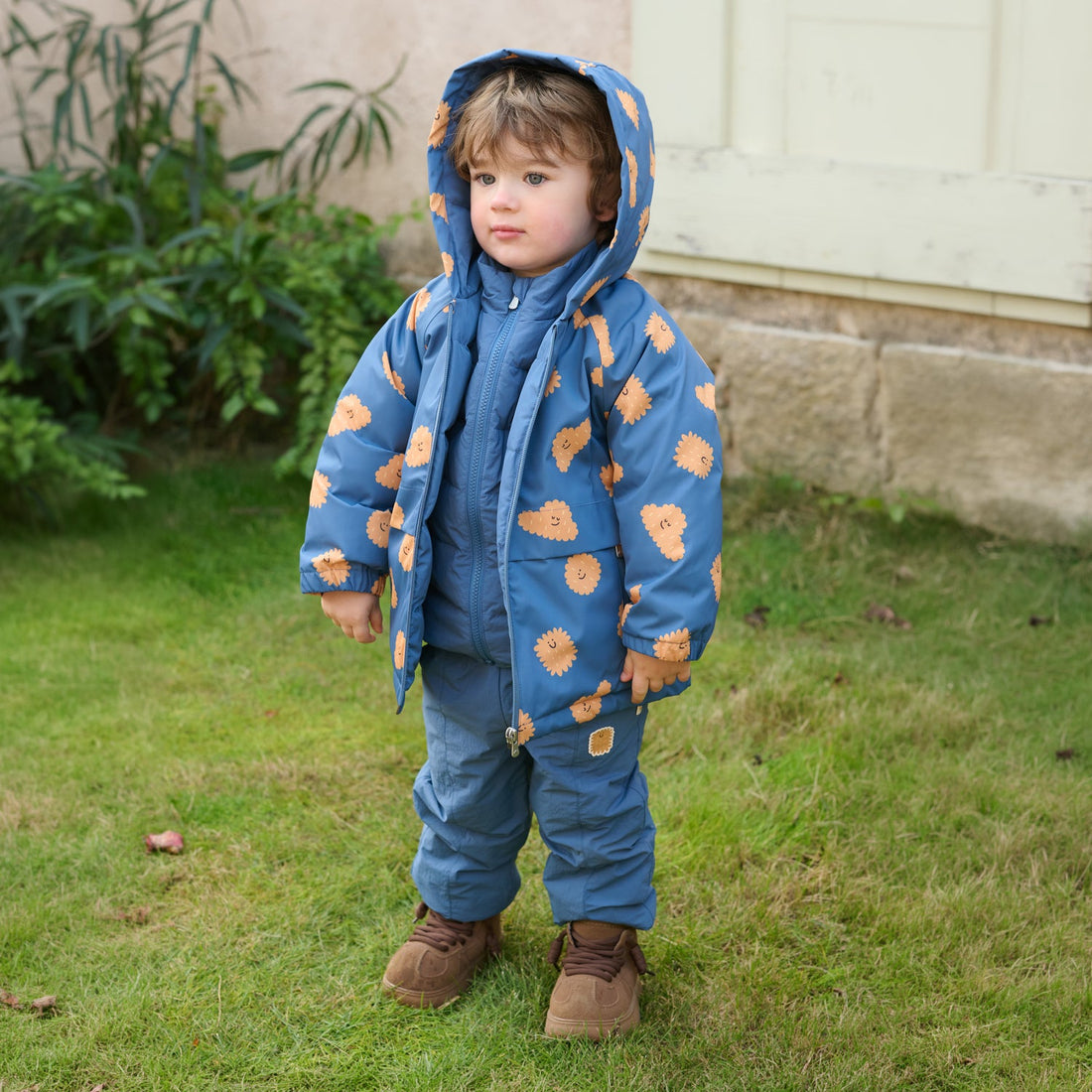 Child wears the Nest Designs Biscuit Banter Waterproof Down Jacket shell hood-up with matching blue pants on grass