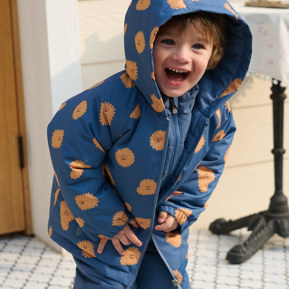 Laughing child bends forward in the Nest Designs Biscuit Banter Waterproof Down Jacket shell with the printed hood raised