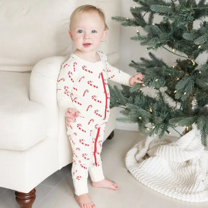 Parade Organic Signature Print 2-Way Zip Matching Holiday Romper, white with red candy cane print, worn by baby beside a Christmas tree.