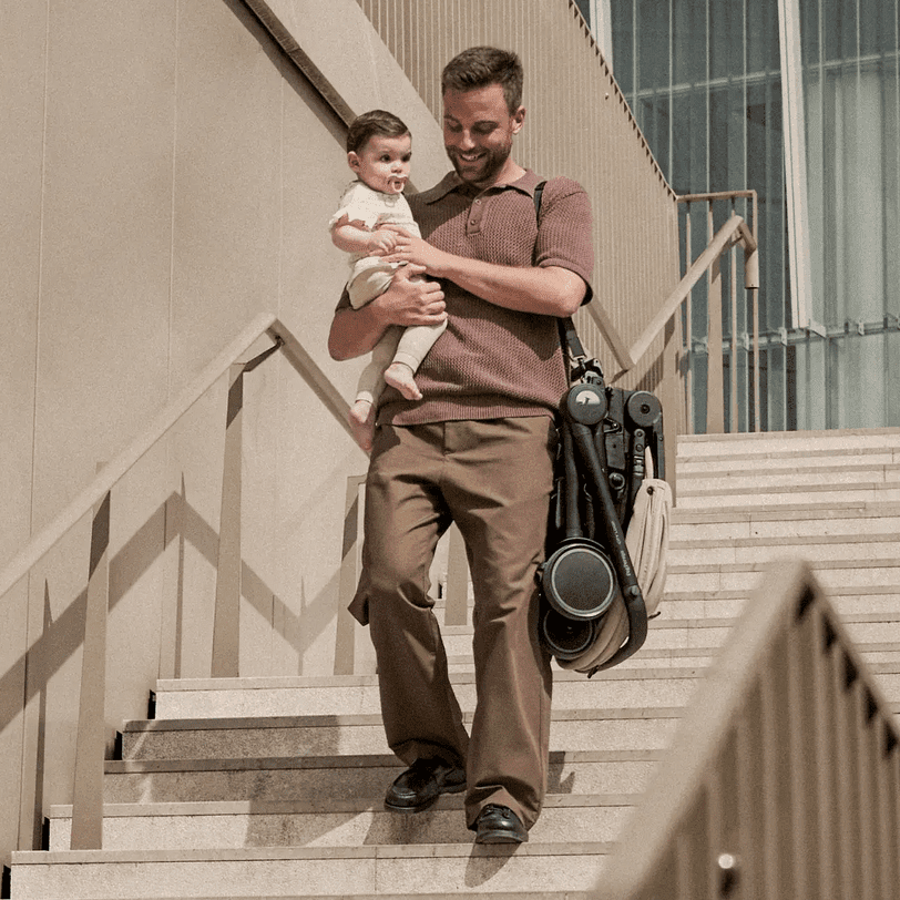 Peg Perego City Loop Stroller Seat compactly folded and strapped to dad’s side as he carries a baby down stairs.
