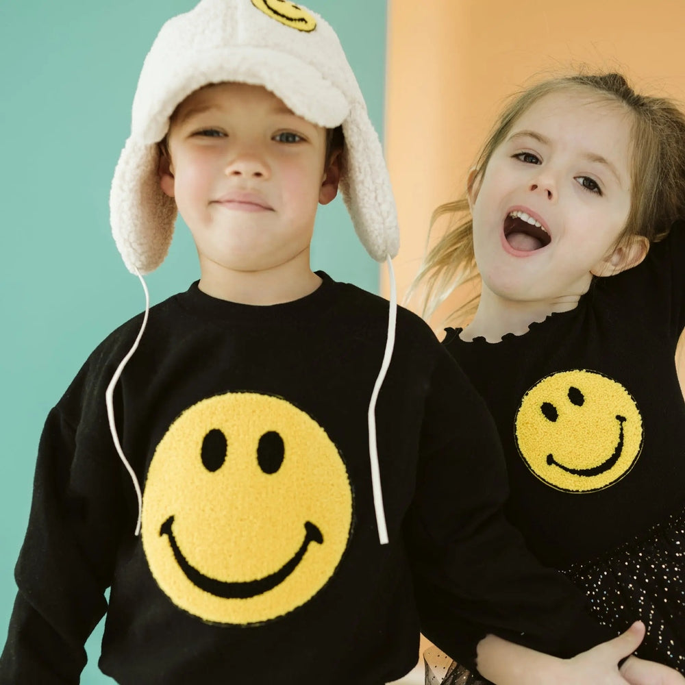 Petite Hailey Smile Smile Sweatshirts Black worn by two kids, big yellow smiley on front; one hugs the other, showing cozy warmth.