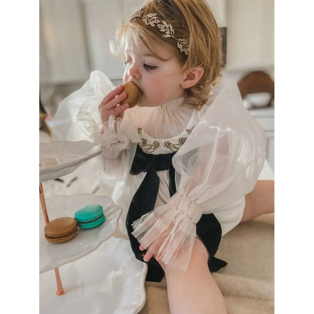 Petite Maison Kids Aurora Ivory Velour Romper with sheer puff sleeves and black bow, a young girl snacks macarons.
