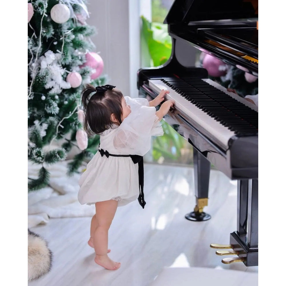 Petite Maison Kids Aurora Ivory Velour Romper worn by a toddler reaching piano keys beside a festive tree.