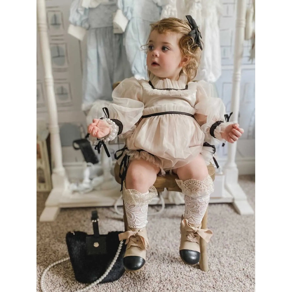 Petit Maison Kids Coco Caramel romper on a baby girl seated on a wooden chair in cream sheer puff sleeves with black trim.