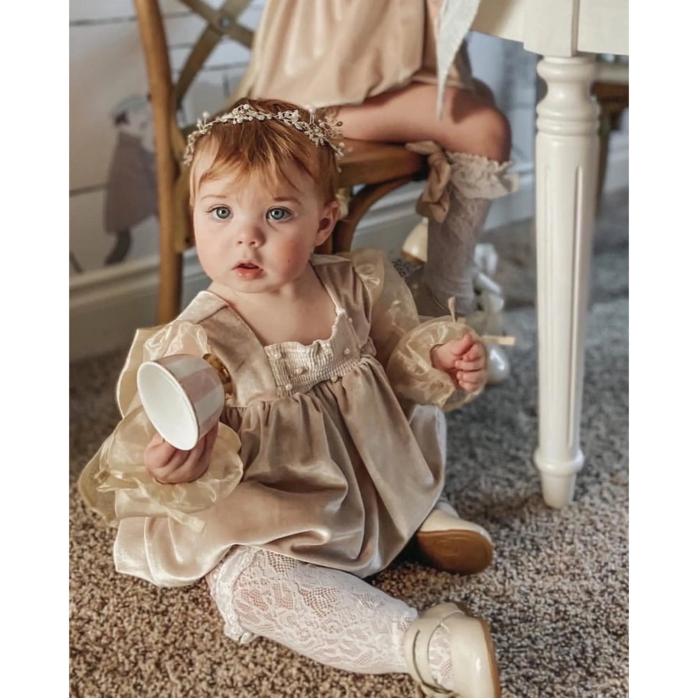 Petite Maison Kids Helena Velour Romper: baby sits on carpet in beige velour with lace sleeves, holding a