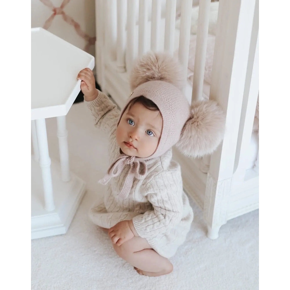 Petite Maison Kids Honeycomb Dusty Pink Cashmere Bonnet on a baby kneeling beside a white crib, fluffy pom-poms.