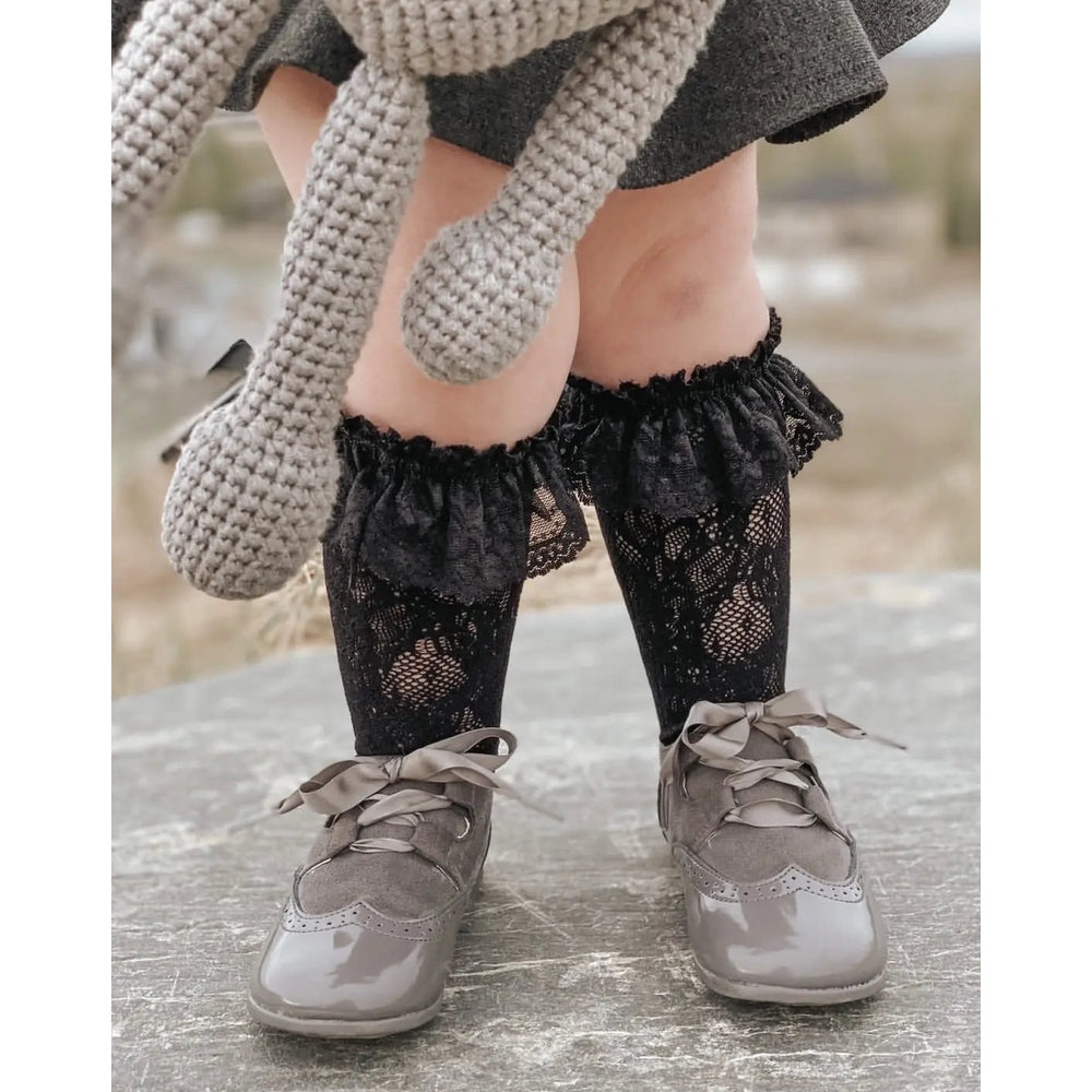 Petite Maison Kids Lola Lace Socks black lace thigh-highs with ruffled top, worn by a child in gray shoes.