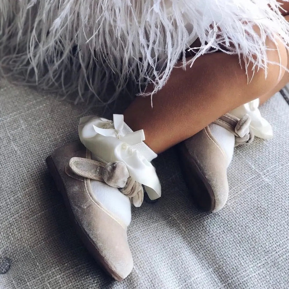 Petite Maison Kids Satin Frilly Socks Ivory with white bows, worn by a child resting on a sofa.
