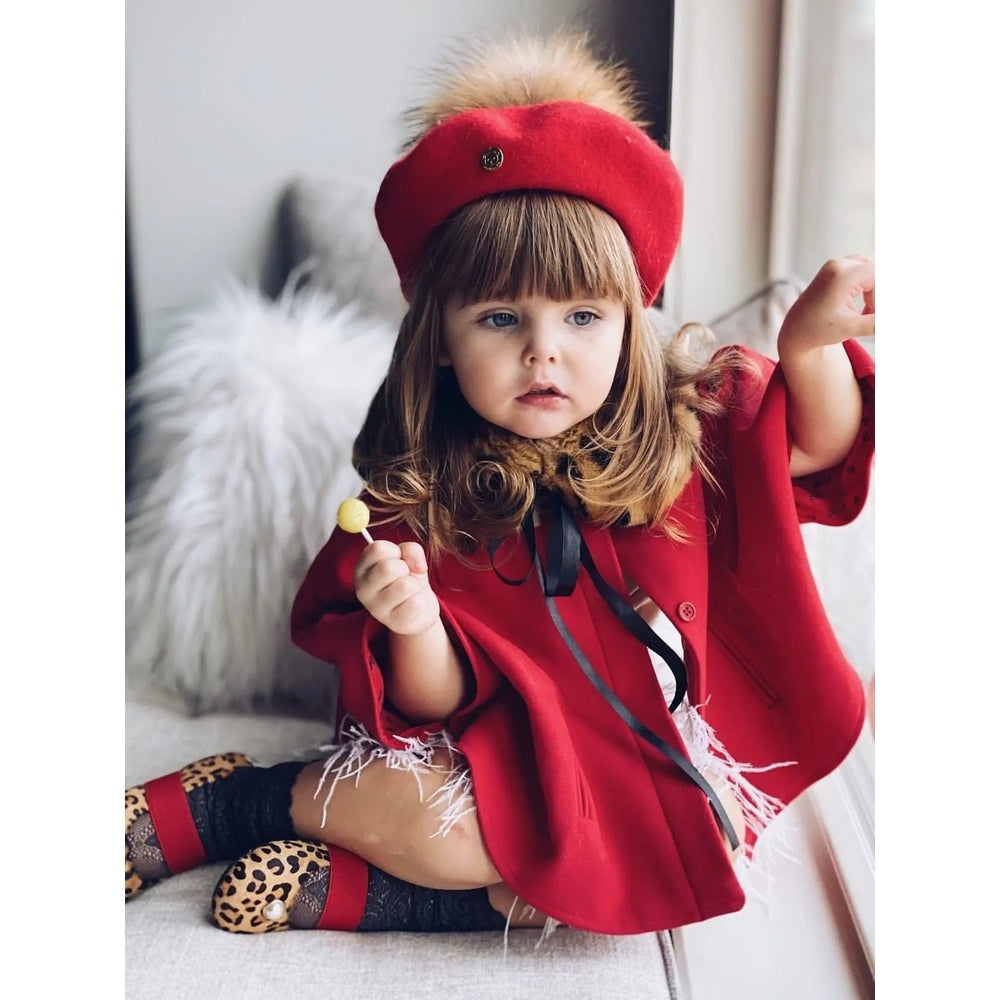 Petite Maison Kids Wool Beret With Pom Pom in red perched on a toddler's head as she holds a lollipop.