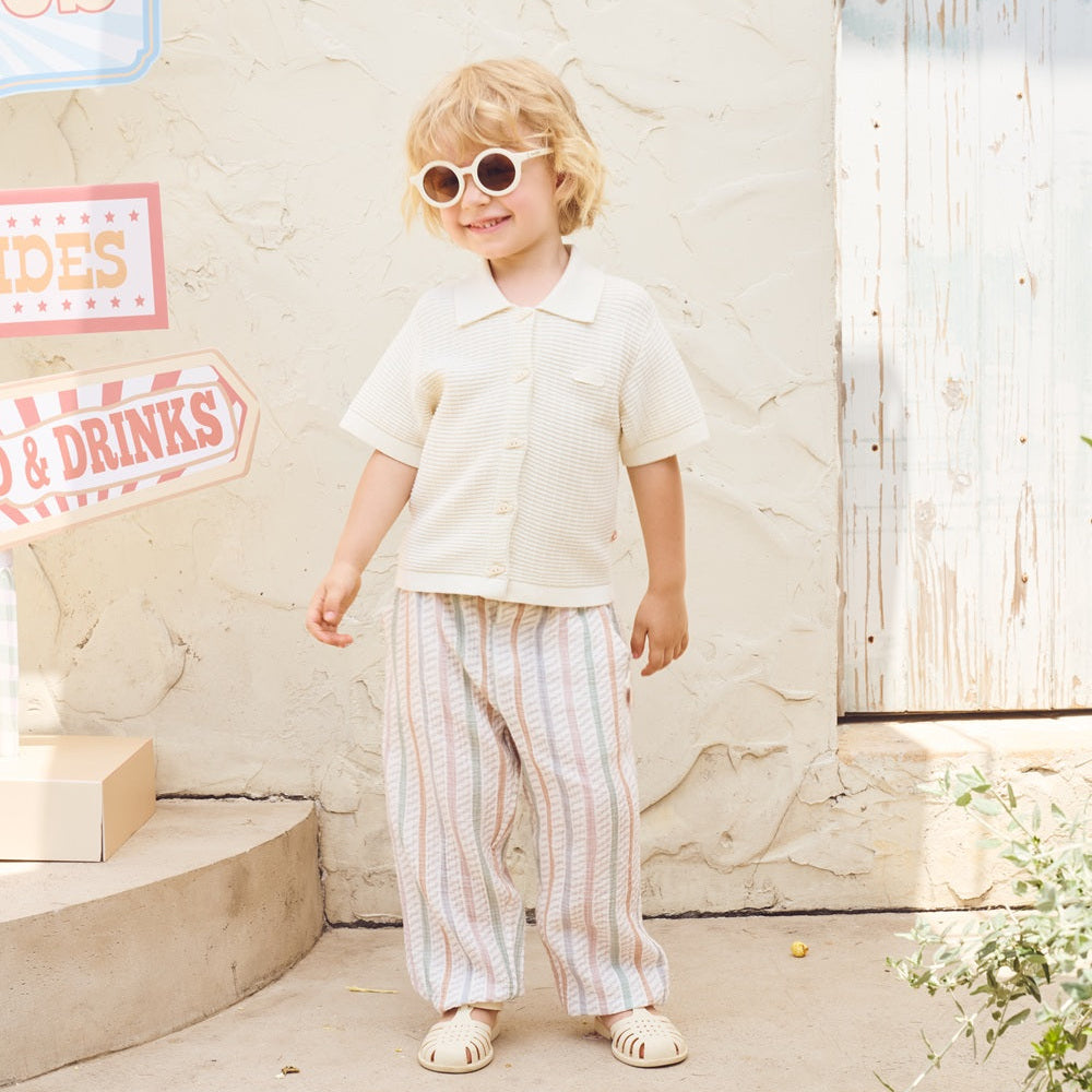 Child wearing sunglasses and a white shirt with striped pants standing in front of a light-colored wall.