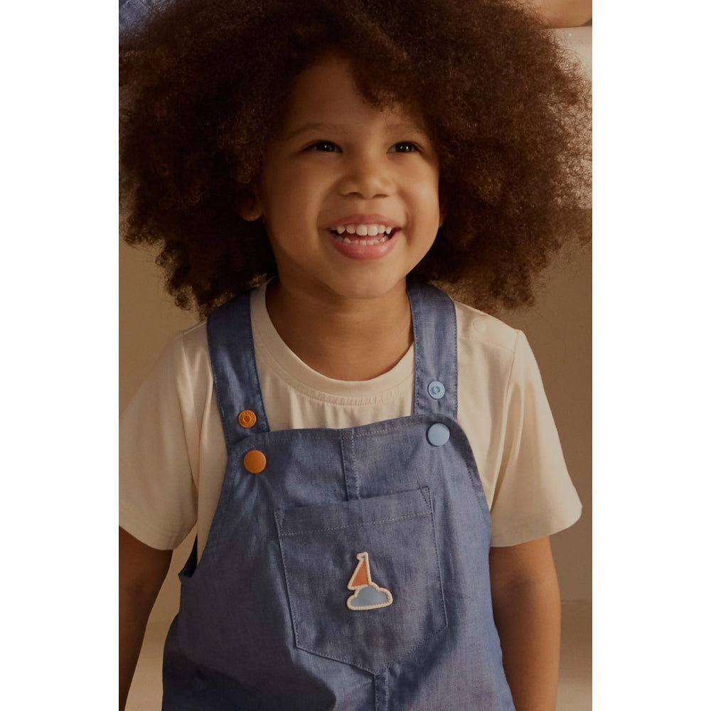 Petite Revery Denim Dress Overalls Sky worn by a smiling toddler in a beige shirt, blue denim with cloud patch on the pocket.