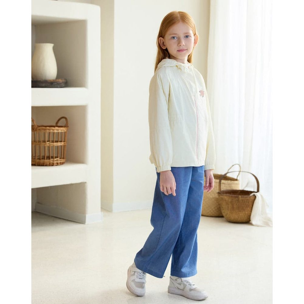 Petite Revery Hooded Windbreaker Jet Stream worn by a girl indoors, cream jacket with zip and hood, paired with blue pants.