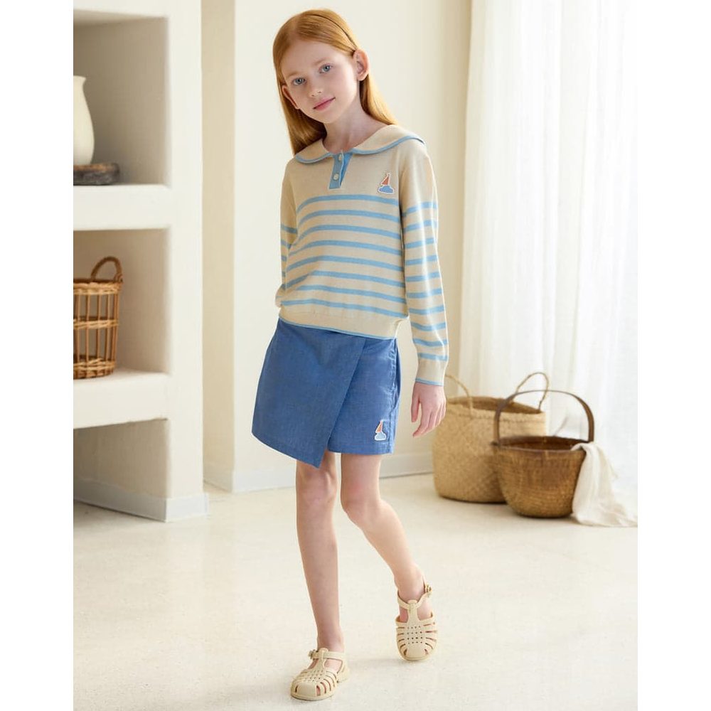 Petite Revery Knit Sweater Seastripes on a toddler girl wearing beige and blue striped knit top with blue shorts, posing indoors.