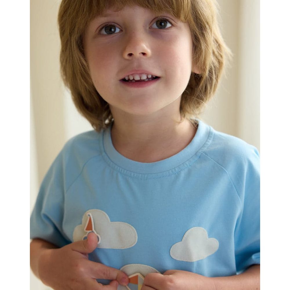 Petite Revery raglan short-sleeve baby tee in Forget-Me-Not blue with cloud patches, child gently adjusting the hem.