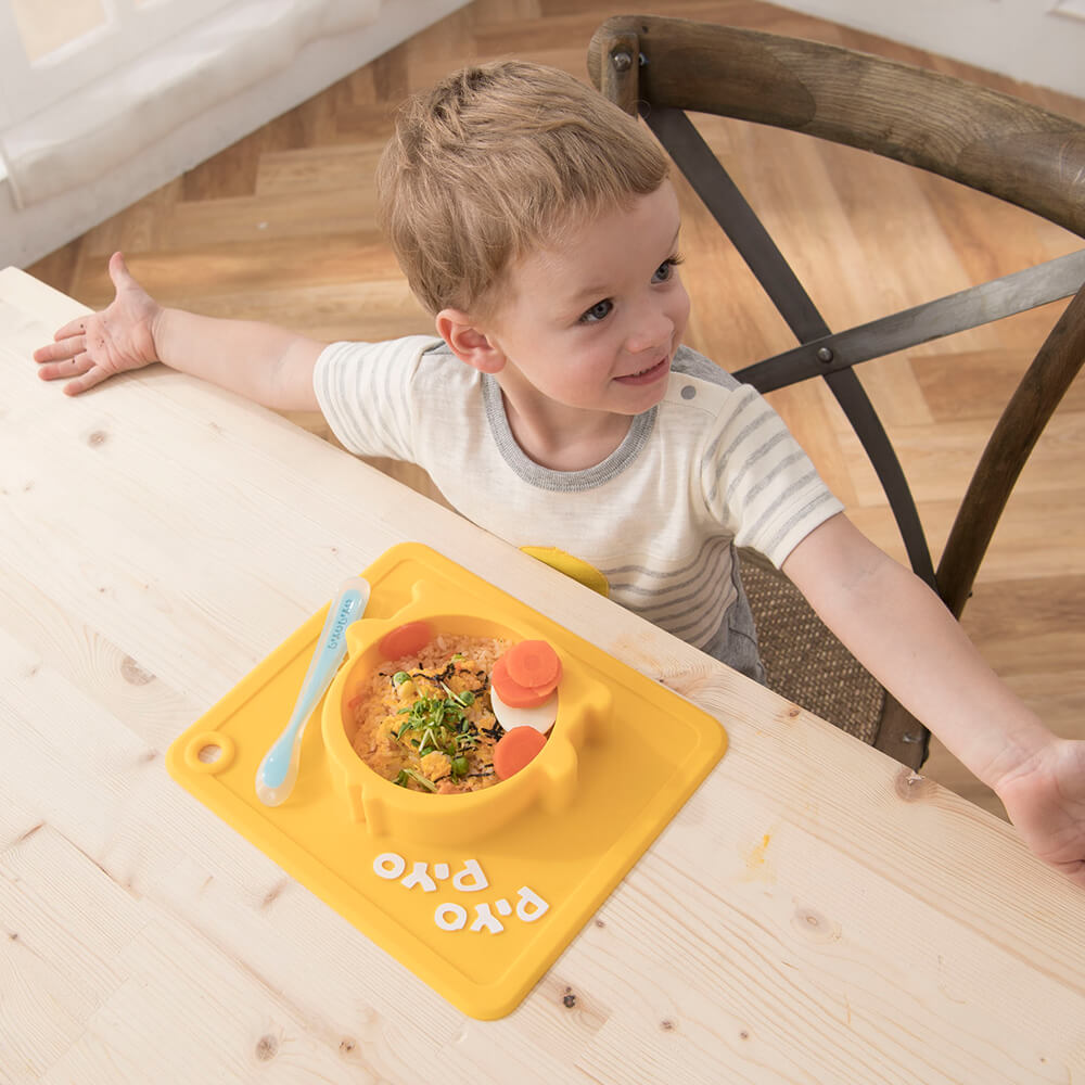 Piyo Piyo anti-slip silicone bowl in yellow suction base with blue spoon; toddler eating at table.