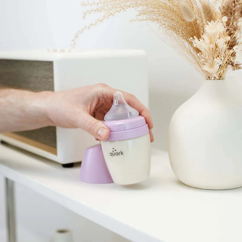 Quark Buubibottle Hybrid Feeding Bundle bottle with lavender lid and silicone nipple, held by a hand on a white shelf.