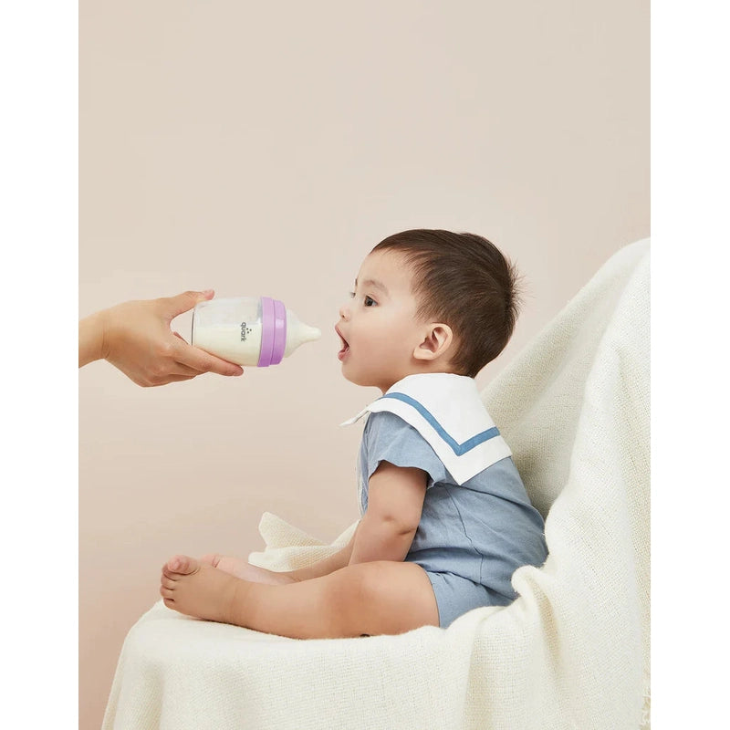 Quark Buubibottle Max Hybrid Feeding Bottle 2 Pack; baby drinks from a white bottle with purple cap held by caregiver.
