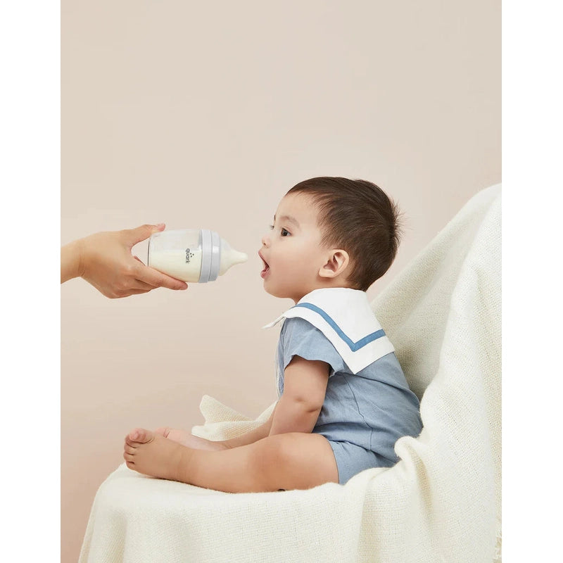 Quark Buubibottle Realfeel bottle nipples; baby in blue romper being fed from a white/gray nipple bottle.