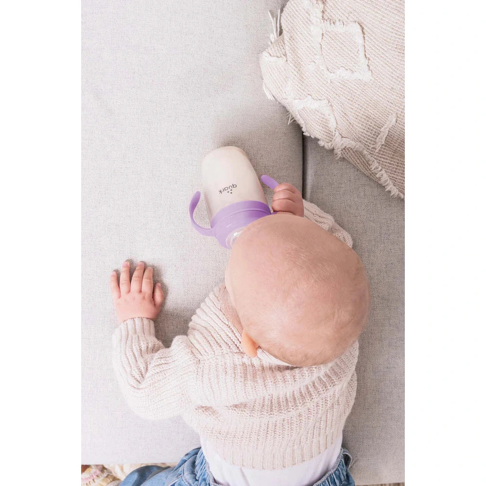 Quark Buubibottle Sip Toddler Sippy Cup With Straw, purple lid, being held by a baby sipping on a sofa.