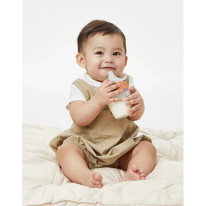 Quark Buubibottle Sip Toddler Sippy Cup With Straw, clear bottle with orange lid, being held and sipped by a smiling baby.