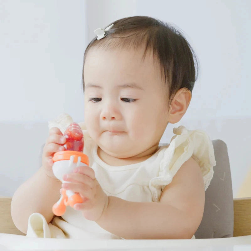 Quark Fruuti baby fruit feeder pacifier with orange handle and white cap held by baby