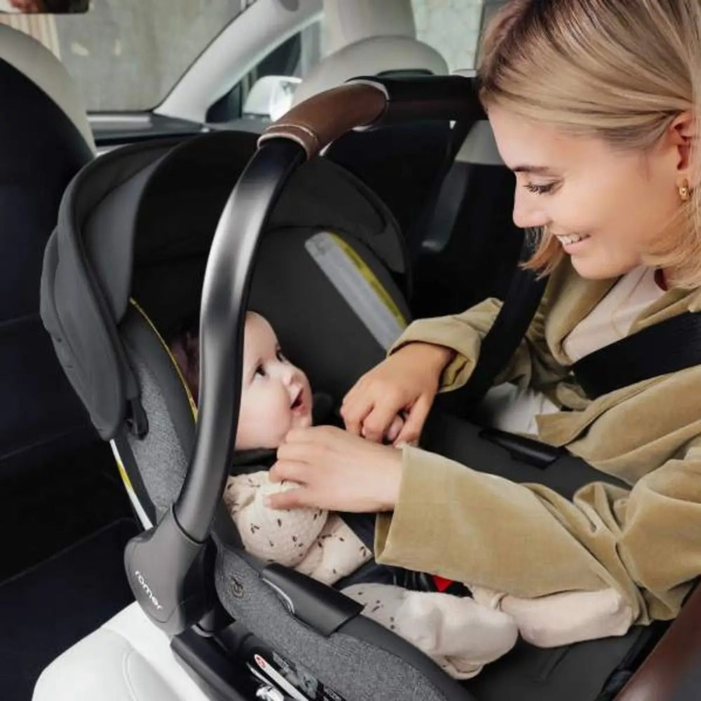 Romer Juni Infant Car Seat with Versa Base in gray fabric, mother buckles the harness on a smiling baby.