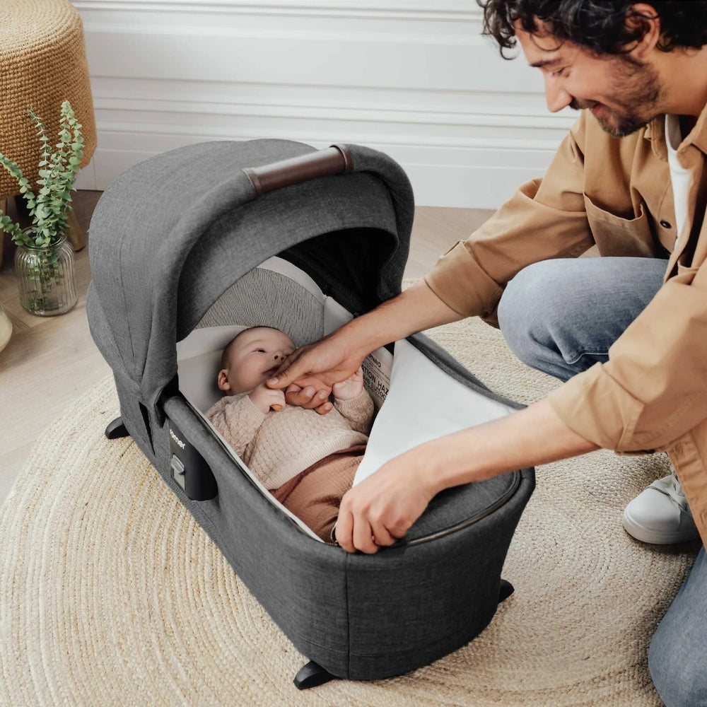 Romer Sera Bassinet for Tura Stroller in gray fabric; parent gently places baby inside the cozy bassinet.