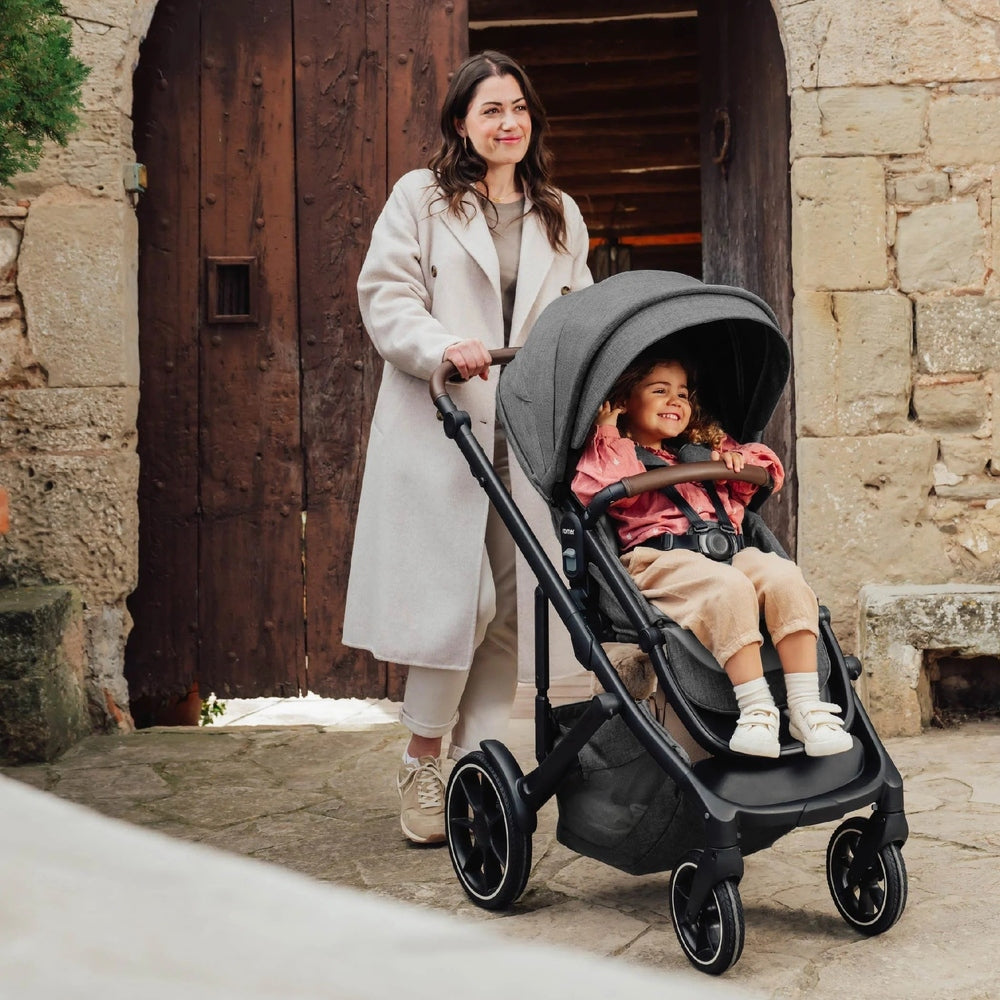 ROMER Romer Tura Stroller in gray pushed by mom outside a rustic doorway, child smiling in full-sized seat.