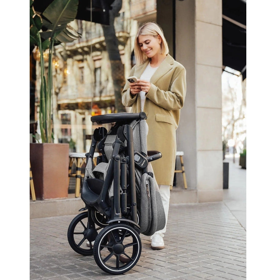 Romer Tura Stroller in grey fabric folded for easy carry, wheels and frame visible as a woman checks her phone.