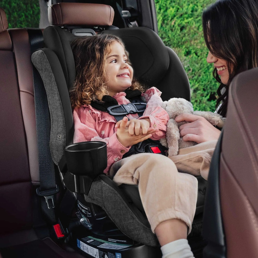Romer Veni Convertible Car Seat, dark gray with five-point harness, child buckled in beside a smiling caregiver.