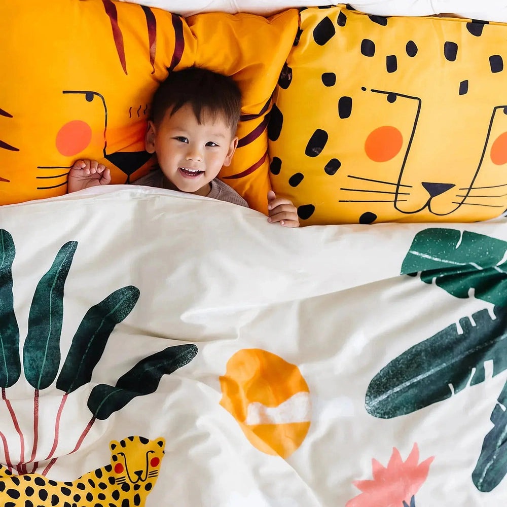 Rookie Humans Kids Bedding Set In The Jungle: child smiles under leafy duvet with orange tiger-print pillowcases.