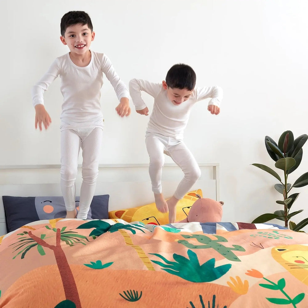 Rookie Humans Kids Bedding Set In The Savanna on a bed; vibrant safari print duvet as two kids jump in white pajamas.