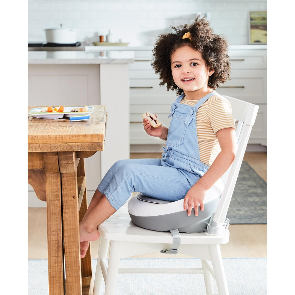 Skip Hop Sleek Seat Booster grey/white; toddler in blue overalls seated at table eating a snack