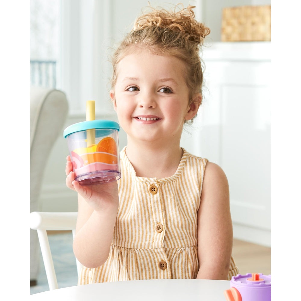 Skip Hop Zoo Shake It Up smoothie cup with turquoise lid and yellow straw, orange slice design, held by smiling child.
