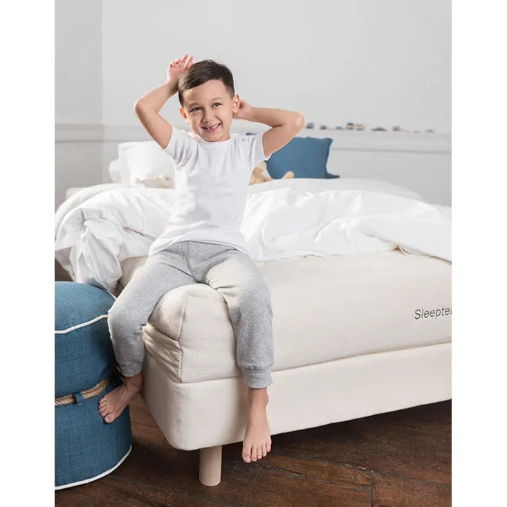 Sleeptek Organic Kids Rubber Mattress with white bedding; a smiling child sits on the edge of the bed.