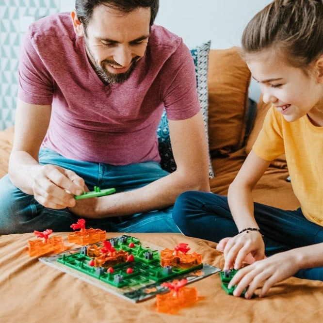 Smart Games Dragon Inferno educational board game; green and orange plastic pieces, family playing on a bed.
