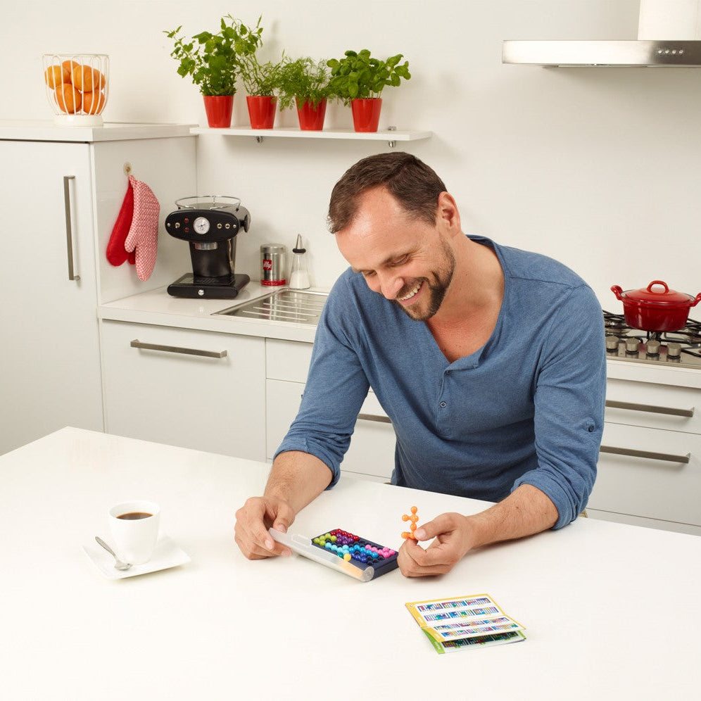 Smart Games IQ Fit puzzle game with colorful pegs on a compact tray; man plays at a white kitchen counter.