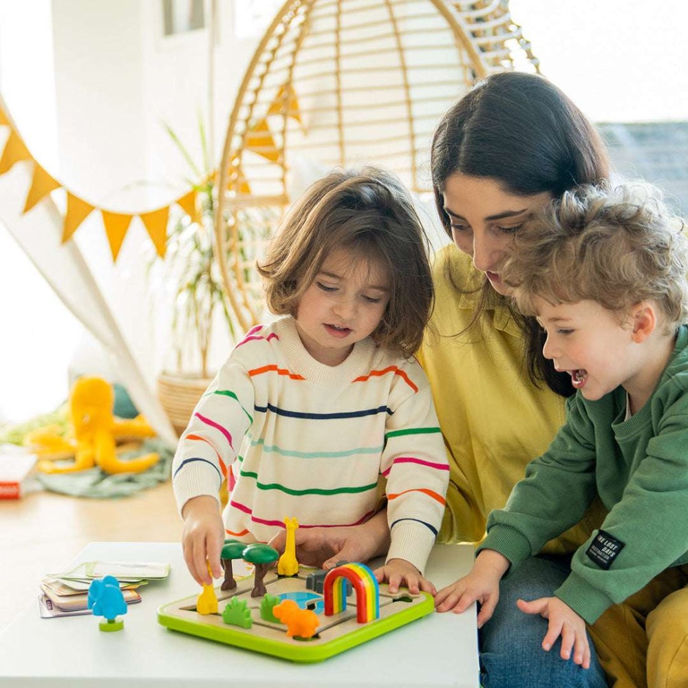 Smart Games Safari Park Jr. educational toy set with colorful animal figures on a green board, played by a mother and two kids.