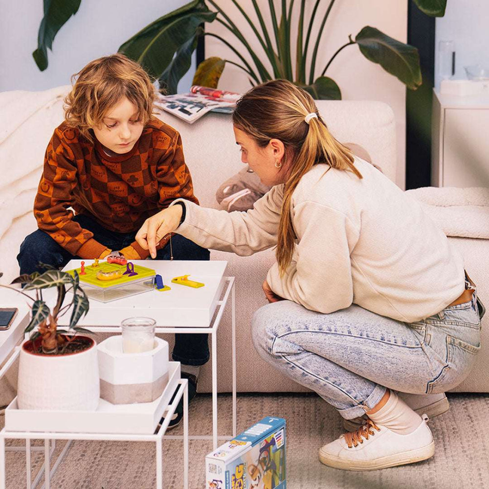 Smart Games Smart Dog Obstacle Run board game; child and caregiver play on a white coffee table with colorful pawns, SMART GAMES.