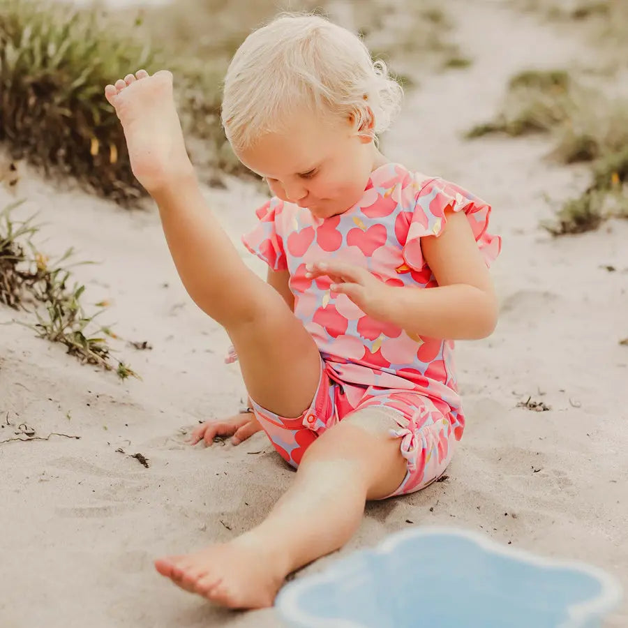 Snapper Rock Apple Love Flutter Sleeve Sunsuit, pink heart print, toddler kicking leg in sand at the beach.