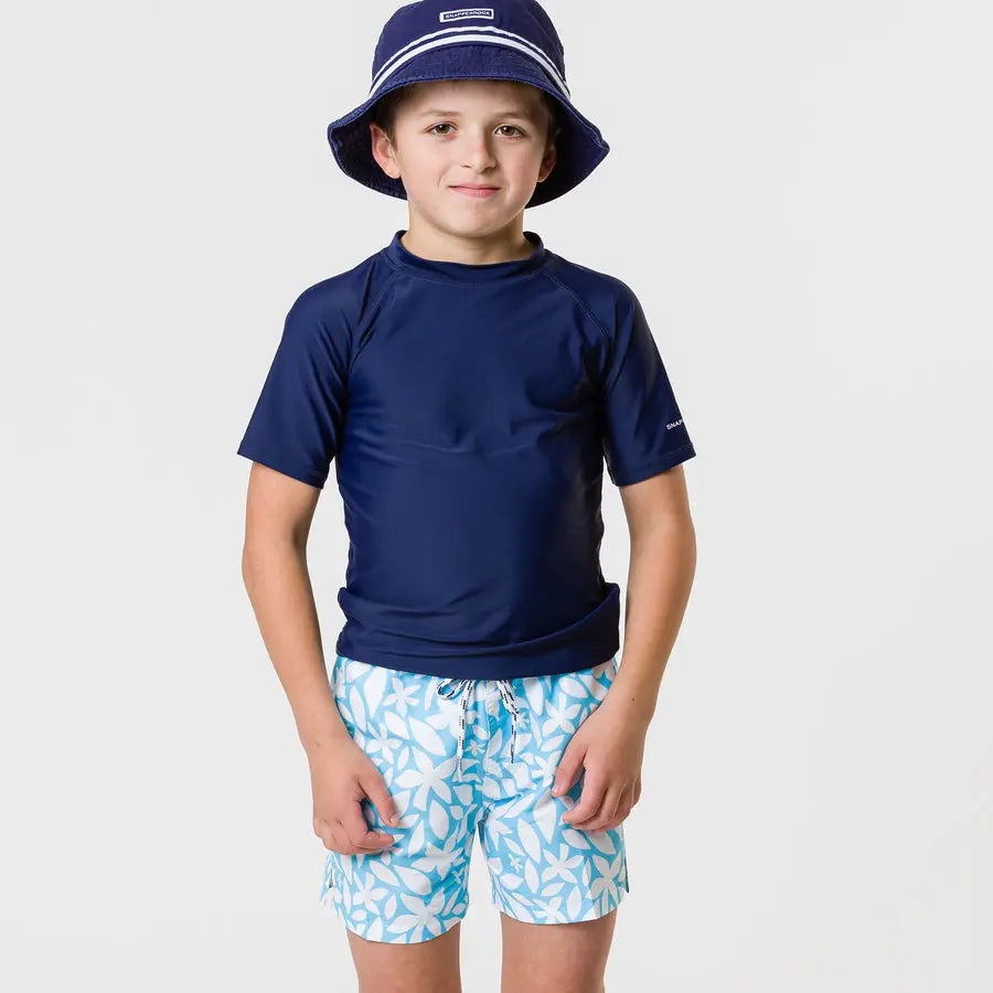 Snapper Rock Aqua Bloom Volley Board Short worn by a boy in a navy rash guard and blue hat, aqua floral print.