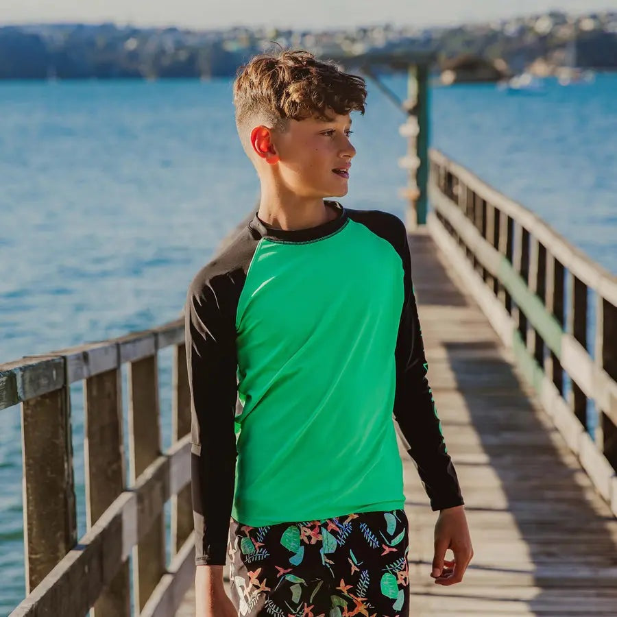 Snapper Rock Green Black Sustainable Ls Rash Top worn by a boy on a pier by the sea, green torso with black sleeves.