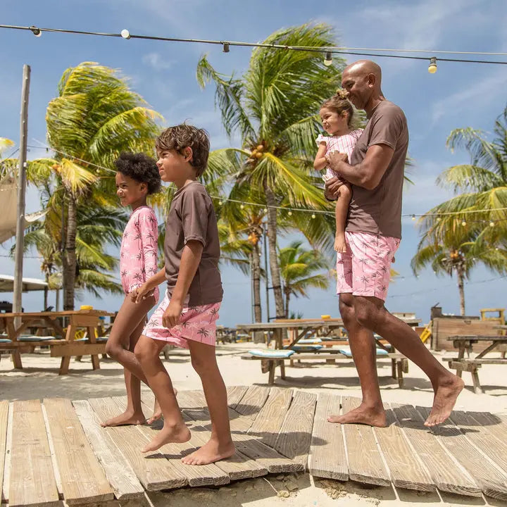 Snapper Rock Palm Paradise Surf Suit in pink palm print; family walks barefoot on a beach boardwalk in matching swimwear.