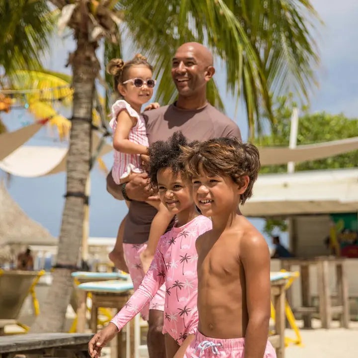 Snapper Rock Palm Paradise Surf Suit; pink palm-print toddler swimsuits worn by smiling kids at a sunny beach.
