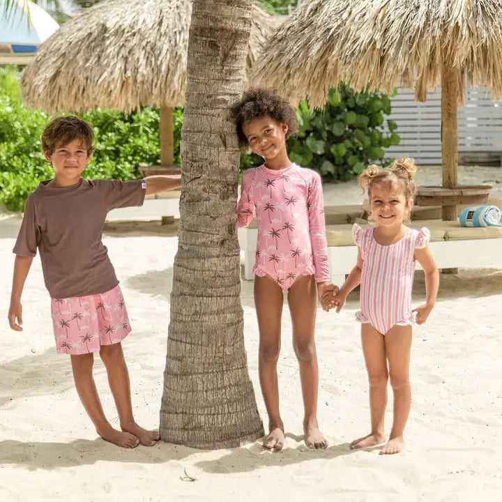 Snapper Rock Palm Paradise Surf Suit trio on a sunny beach, kids in pink palm-print rash guard and striped swimsuits, holding hands.
