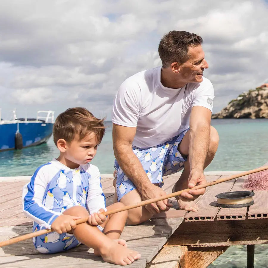 Snapper Rock Penguin Patrol LS Sunsuit; toddler in blue penguin print reels a rod with dad on a sunny dock.