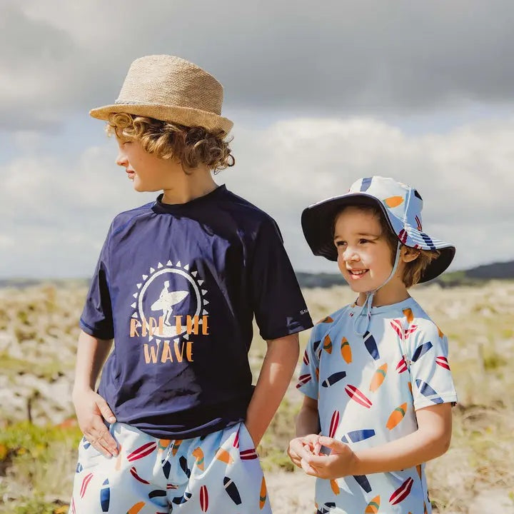 Snapper Rock Retro Surf Baby Set: navy sun-logo tee and surf-print shorts worn by two kids posing on a sunny beach.